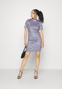 Vestido de manga curta, listrado, com várias tonalidades de roxo, rosa e branco. Combinado com sapatos de salto pretos e uma pequena bolsa preta.