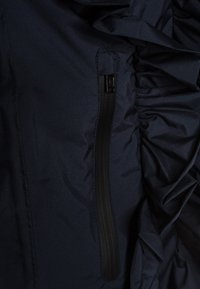 Námořnická bunda s boční kapsou na zip a řasenými akcenty. Materiál je hladký s mírně lesklým povrchem. Viditelné jsou i detaily švů.