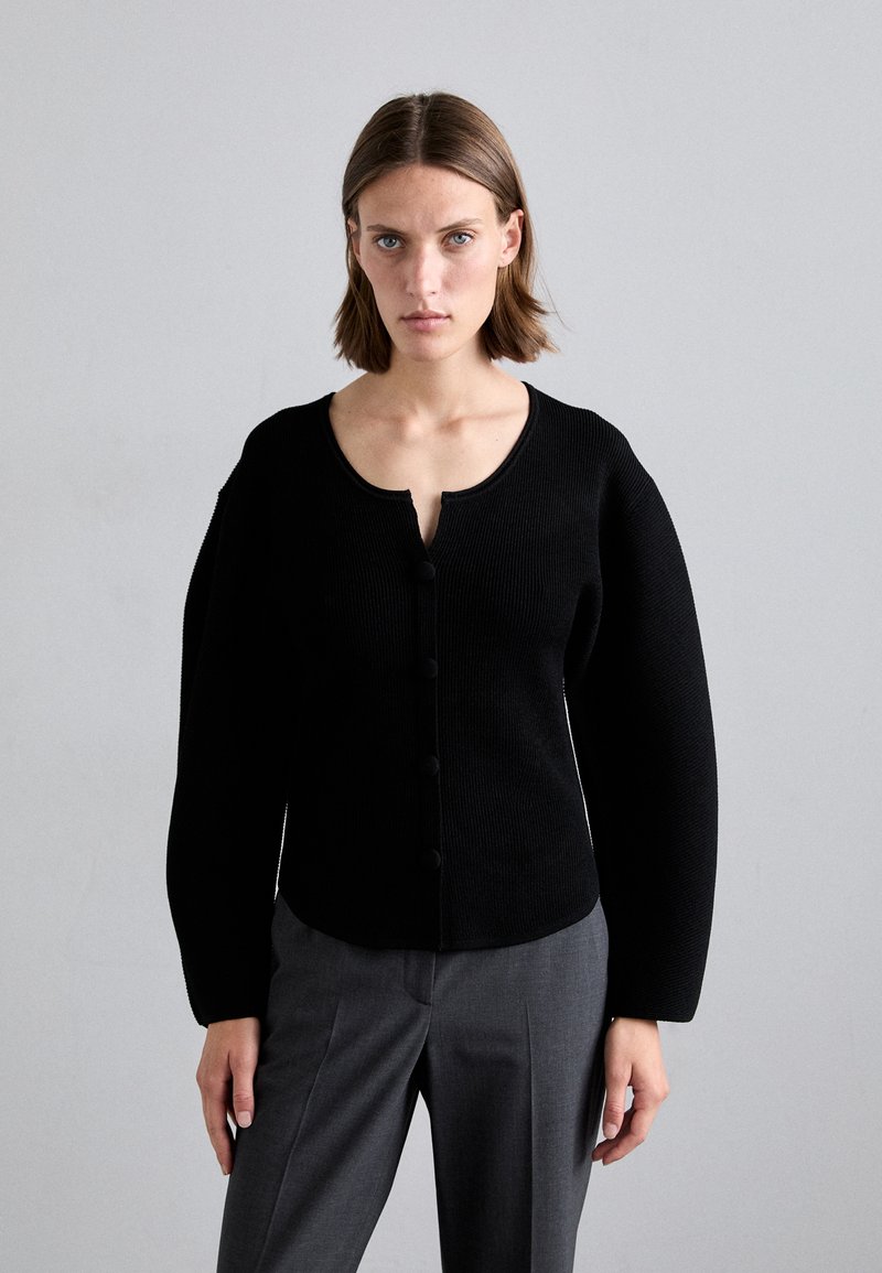 Black ribbed cardigan with a round neckline, button front closure, and exaggerated sleeves, paired with gray trousers.