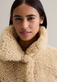 Une jeune femme aux cheveux bruns porte un épais manteau en polaire crémeux et bouclé avec un large col sur un fond neutre.
