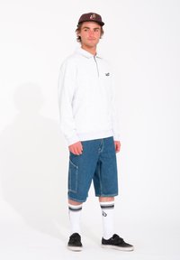 Suéter de manga larga gris claro con cuello de cremallera y logo, combinado con pantalones cortos de mezclilla azul hasta la rodilla, calcetines blancos a rayas y zapatos negros.