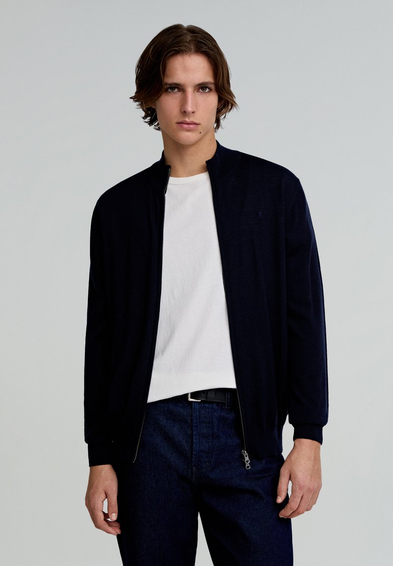 Geacă navy cu fermoar și guler în picioare, realizată dintr-un material texturat, asortată cu un tricou alb cu guler rotund și pantaloni din denim închisi.