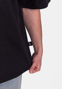 Chemise noire à manches courtes présentée avec une étiquette latérale. Tissu en coton, coupe décontractée, couture visible et texture lisse ; main reposant sur l'ourlet.