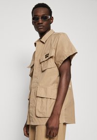 Beige short-sleeve shirt with four pockets and a front button closure. Fabric appears lightweight with a textured finish. Black logo patch on chest.