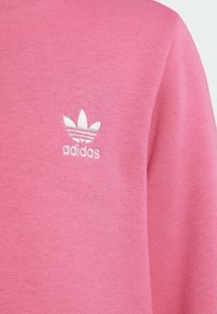Ružová mikina vyrobená z jemného materiálu, s bielym vyšívaným logom Adidas, ktoré má tri listovité akcenty a horizontálne pruhy.