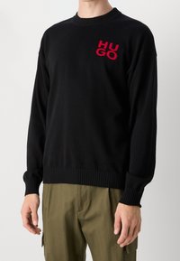Pull noir en maille avec encolure ronde, arborant un logo brodé rouge "HUGO" sur la poitrine gauche. Poignets et ourlet côtelés.