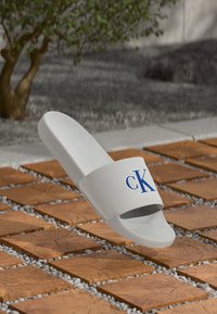 Fehér csúszó szandál kék "cK" logóval a pánton, faforgácsból készült négyzetes lapok felett, kis fehér kövekkel egy szabadtéri környezetben.