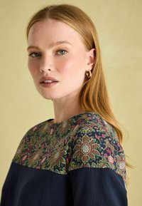 Navyblaues Oberteil mit floral gemustertem Schulterbereich. Das Material zeigt ein kunstvolles Design in Grün, Rot und Blau. Rundhalsausschnitt und lange Ärmel.