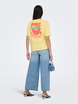 Kvinde iført en gul T-shirt med "Pink Grapefruit"-tryk, vide blå jeans, teal kitten heels og holder en lille blå håndtaske.