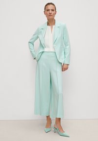 Mintgroene, op maat gemaakte blazer met een enkele knoop, bijpassende wijde broek en een witte top met een sleutelgat-halslijn. Mintgroene hakken maken de outfit compleet.