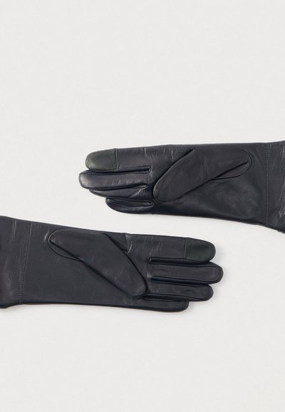 Μαύρα δερμάτινα γάντια με λεία υφή, διακοσμημένα με πράσινη πινελιά στις άκρες των δαχτύλων και αεροδυναμικό σχεδιασμό. Δύο γάντια απλωμένα επίπεδα.