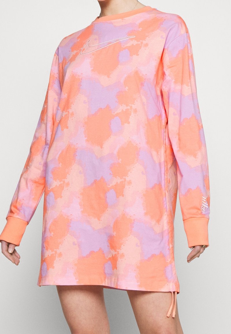 Dlouhé rukávy, oversized šaty v oranžovém a lila tie-dye vzoru. Mají kulatý výstřih a vyšívané logo Nike v stříbrné barvě.