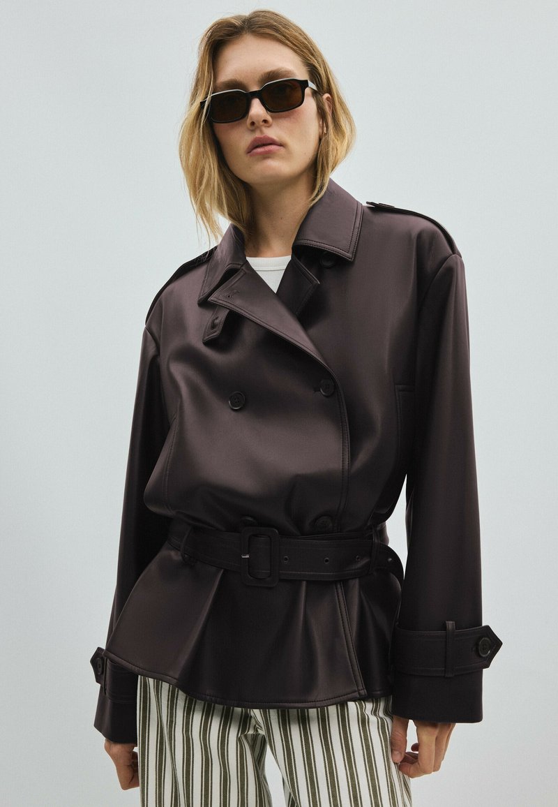 Femme portant une veste en cuir noire ceinturée, un pantalon rayé et des lunettes de soleil rectangulaires foncées sur un fond clair uni.