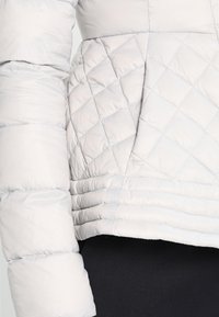 Chaqueta acolchada gris claro con patrón acolchado en el cuerpo, bajo acanalado y textura de tela lisa y brillante.