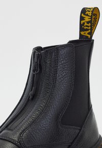 Bottine en cuir noir avec fermeture éclair frontale, panneau latéral élastique et languette jaune et noire affichant le nom de la marque.