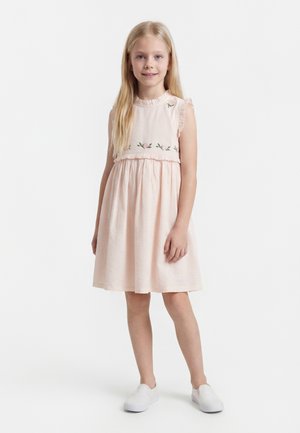 Mladé dievča s dlhými blond vlasmi v bezrukávových bledoružových šatách s kvetinovou výšivkou a bielych nazúvacích topánkach, stojí a usmieva sa.