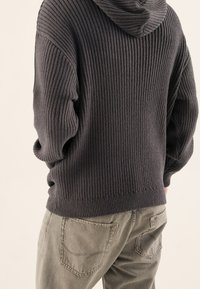Dunkelgrauer Rippstrick-Hoodie mit Kapuze und lässigem Schnitt. Kombiniert mit hellen, strukturierten grünen Hosen mit hinteren Taschen.
