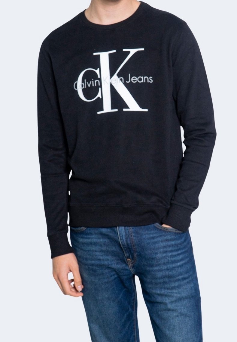 Persona che indossa una felpa nera a maniche lunghe Calvin Klein Jeans con un grande logo bianco "cK" e jeans blu, in piedi con una mano in tasca.