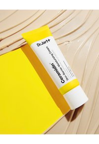 Dr. Jart+ Ceramidin hudbarrier fuktighetskremtube har en hvit kropp med et gult lokk, plassert på en teksturert beige bakgrunn.