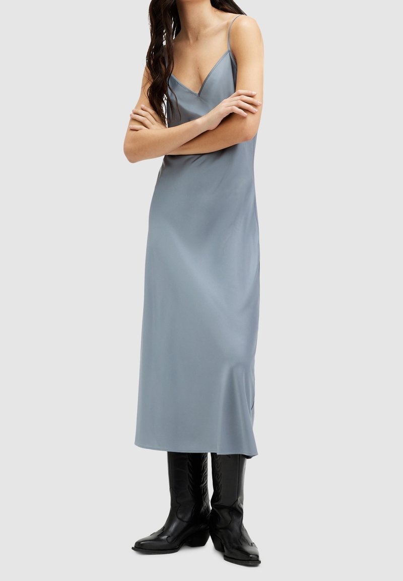 Femme portant une robe midi sans manches bleu clair avec un décolleté en V et des bottes noires montant jusqu'aux genoux, debout les bras croisés.