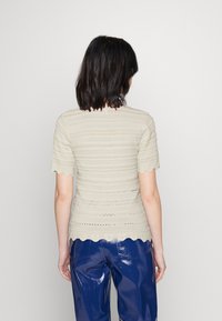 Pletený svetr v světle béžovém odstínu, s texturovaným vzorovaným designem, ozdobený vroubkovanými okraji a krátkými rukávy. Kombinován s lesklými modrými kalhotami.