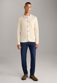 Cardigan beige testurizzato con colletto e tasca frontale, chiusura a bottoni; abbinato a jeans blu scuro e stivali marroni alla caviglia. Sfondo neutro.