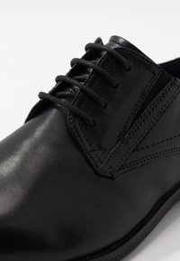 Scarpa in pelle nera con superficie liscia, design con lacci e dettagli cuciti. La punta è arrotondata, con una leggera lucentezza.