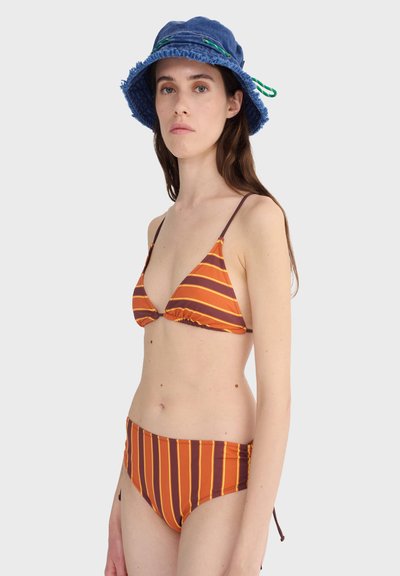 Femme portant un bikini rayé orange et marron à fines bretelles et un chapeau seau bleu avec des détails verts, debout devant un fond clair uni.
