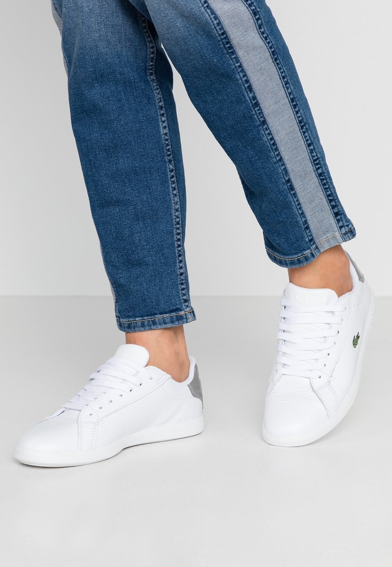 Pieds portant des baskets en cuir blanc avec des accents gris, associées à un jean bleu avec une bande latérale gris clair.