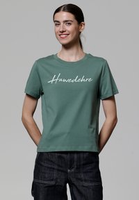 Grünes Baumwoll-T-Shirt mit kurzen Ärmeln und Rundhalsausschnitt. Die Vorderseite zeigt den weißen Schreibschrift-Text "Hawedehre." Schlichter Rücken und lockere Passform.