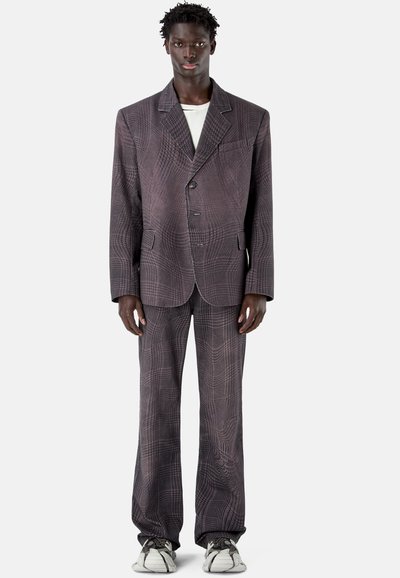 Hombre de pie, vestido con un traje ancho de patrón gris oscuro, pantalones anchos y zapatillas, fondo claro y liso.