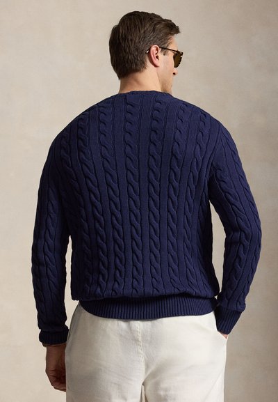 Muž v tmavomodrom vrkočiakom pulóvri a bielych nohaviciach, zobrazený zozadu, s okuliarmi na slnko a krátkymi hnedými vlasmi.