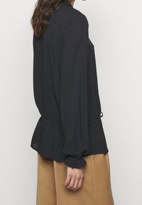 Černá halenka s dlouhým rukávem, která má peplum lem a stažené manžety, vyrobená z lehkého materiálu s hladkou texturou.