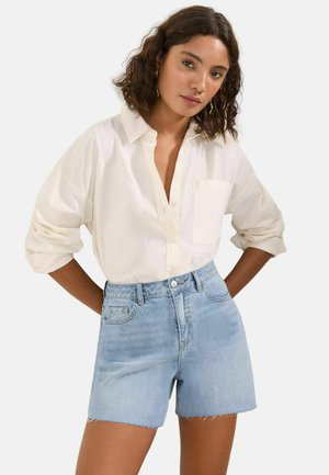 Göndör hajú nő, aki krémszínű hosszú ujjú inget visel felhajtott ujjakkal és világoskék, magas derekú farmer rövidnadrágot.