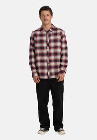 Camicia in flanella a quadri rossi e blu, con bottoni frontali, maniche lunghe e un piccolo logo. Abbinata a pantaloni neri e sneaker casual.