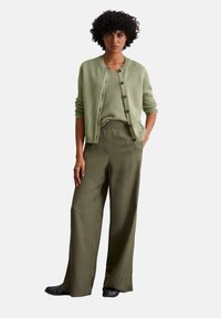 Hellgrüner Strickcardigan über einem passenden Top, kombiniert mit lockeren olivgrünen Hosen und schwarzen Ballerinas.