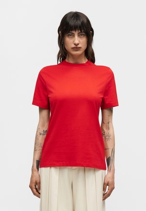 Femme aux cheveux foncés portant une chemise rouge à manches courtes et un pantalon plissé beige, debout devant un fond clair uni.