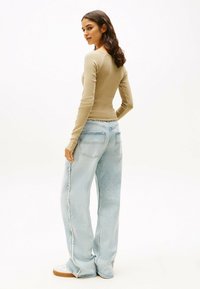Langärmliges, tailliertes beige Oberteil mit gewelltem Saum, kombiniert mit hellblauen, weiten Jeans mit ausgewaschenem Look und zwei hinteren Taschen.