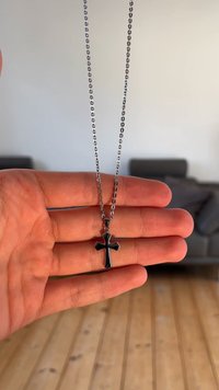 Croce pendant in metallo nero su una collana d'argento tenuta in mano. La croce presenta una finitura lucida e estremità appuntite, con un design della catena semplice.