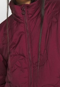 Burgundyröd puffjacka med hög krage, prydd med broderade bladmönster och en central dragkedja. Tillverkad av lätt, strukturerad tyg.