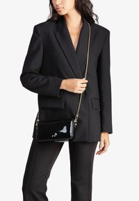 Borsa a tracolla in vernice nera con catena dorata, texture liscia, forma rettangolare e dettagli del logo discreti sul davanti.