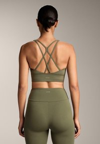 Olivno zelena športna modrček z vzporednimi hrbtnimi trakovi, izdelan iz raztegljivega materiala. Kombiniran z visokim pasom usklajenih legic.