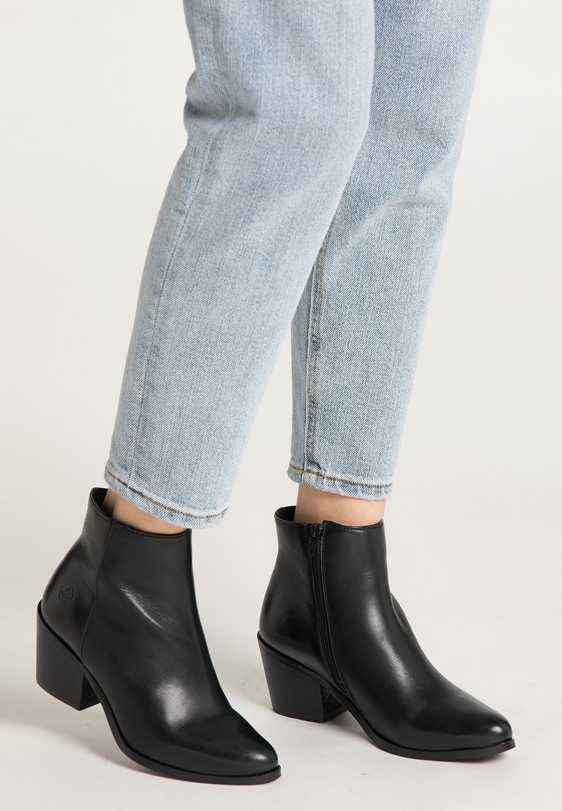 Personne portant un jean bleu clair et des bottines en cuir noir à talons carrés bas, debout sur un fond clair uni.