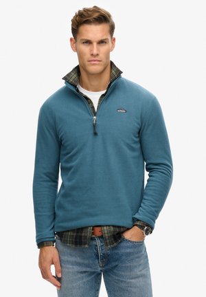 Fleece pullover in teal met een ritskraag, gedragen over een geruit shirt. Bevat een logopatch en geribde manchetten. Gestyled met denim en een horloge.