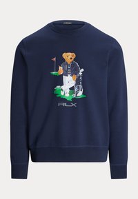 Marinblå sweatshirt med en björn i golfkläder, randig skjorta och jacka, med en golfväska, framhävd av en bakgrund med grön gräsmatta.