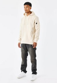 Sudadera beige con capucha, con un bolsillo frontal y una parche negro en la manga, combinada con unos pantalones cargo oscuros y desgastados y zapatillas blancas.