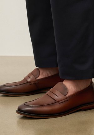 Brūnas ādas loaferes ar reljefa augšu, valkātas kopā ar melnām saīsinātām biksēm uz bēša fona.