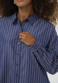 Marineblauwe button-up shirt met witte verticale strepen en kleine stippen, voorzien van een klassieke kraag en omslagmouwen.