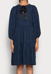 Navy- und blaue Karokleid mit rundem Halsausschnitt, Puffärmeln und einer schwarzen Satinschleife an der Vorderseite. Hat ein gestuftes Rockdesign.
