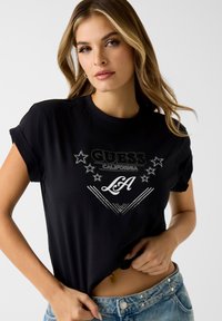 Czarna koszulka z podwiniętymi rękawami, ozdobiona błyszczącym nadrukiem z napisem "GUESS CALIFORNIA" i gwiazdkami, zestawiona z jasnoniebieskimi dżinsami.